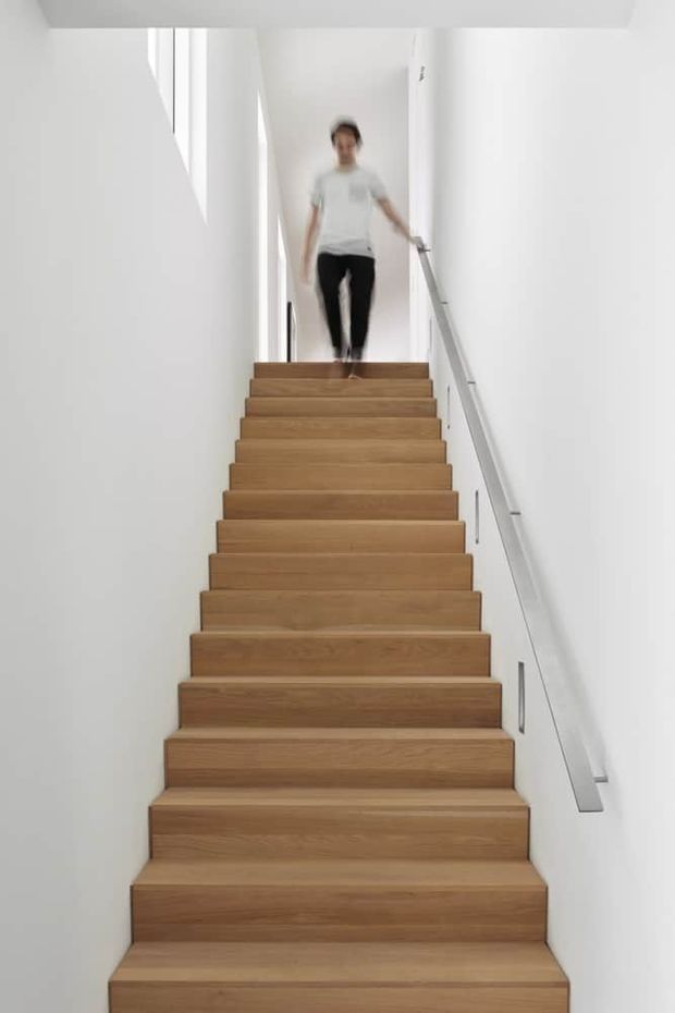 A straight, enclosed staircase with broad solid timber treads and risers ascending through a narrow white-painted stairwell. A simple metal handrail with clear glass balustrade panels runs along the right wall, while vertical glazing or a slim window opening brings in daylight at the upper landing. A person is descending the stairs.
