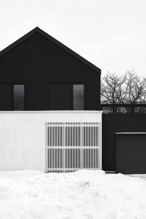 Exterior view of a dark, modern gable-roofed house in a snowy setting. The facade combines vertically ribbed dark cladding with horizontal dark siding, narrow windows, a white slatted privacy screen or balcony enclosure, and a dark garage door. Bare trees and deep snow reinforce the wintry exterior context.