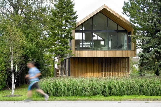 Exterior view of a compact contemporary timber-clad house set among mature greenery. The upper level has a prominent front-facing gabled roof and extensive dark-framed glazing, while the lower level features vertical timber screening and a recessed entry or garage-like opening. Ornamental grasses and trees soften the facade, with a blurred pedestrian in the foreground.