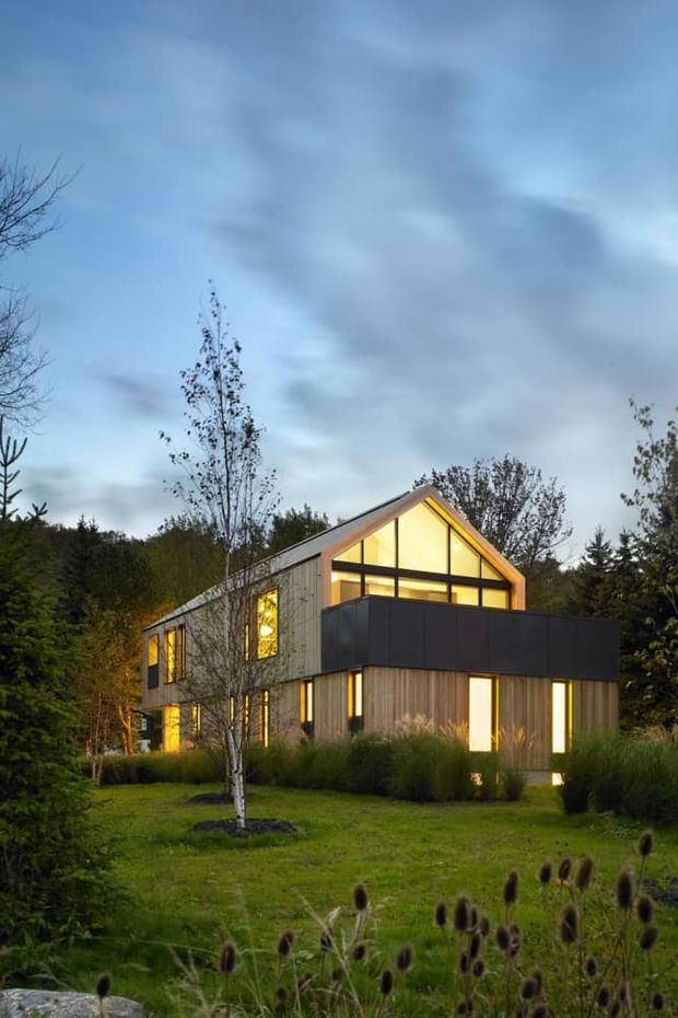 A contemporary two-storey house photographed at dusk in a landscaped rural or wooded setting. The home has a steep gabled roof, vertical timber cladding, extensive warm-lit glazing, and a prominent dark projecting upper-level balcony or box volume. Ornamental grasses and young trees surround the building.