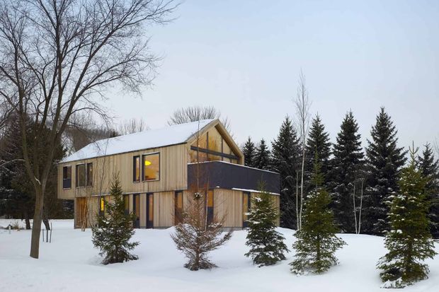 A modern two-storey timber-clad house set in a snowy wooded landscape. The home has a steep gabled roof, large vertical windows, a projecting dark horizontal upper-level volume, and a simple natural-material facade. Evergreen trees and snow surround the building.