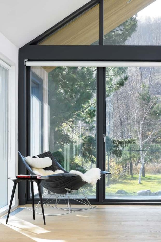 Bright modern interior corner with expansive floor-to-ceiling glazing, black-framed windows, and a triangular clerestory-like upper window beneath a sloped timber-lined ceiling. A sculptural black lounge chair with light cushions and a small side table sit on pale timber flooring, with trees and garden visible outside.