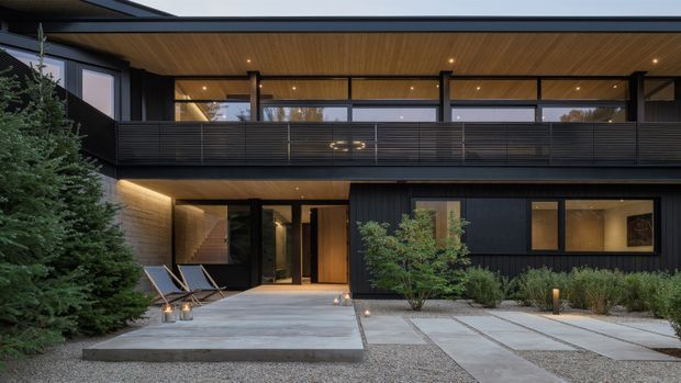Contemporary two-storey residential facade viewed from the front, featuring dark horizontal cladding, extensive glazing, a broad upper-level balcony, warm timber soffits, and a covered entry courtyard. Concrete stepping slabs, gravel landscaping, low planting, and integrated exterior lighting create a refined modern approach.