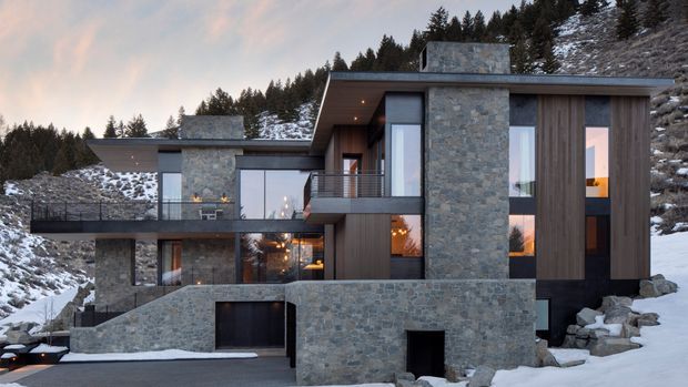 A contemporary multi-level mountain home set into a snowy, forested hillside. The exterior combines extensive irregular natural stone walls with dark vertical timber cladding, broad flat roof planes, large floor-to-ceiling windows, balconies, and a partially concealed garage. Warm interior lighting contrasts with the cool alpine setting.