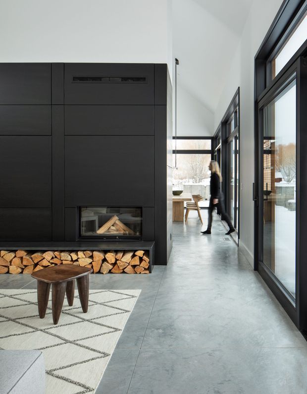 A contemporary open-plan interior centered on a dramatic full-height matte black fireplace wall with integrated firebox and neatly stacked firewood storage. A polished grey concrete floor extends through a glazed corridor toward a dining area, while large dark-framed windows and doors bring in abundant natural light. A patterned rug and sculptural timber coffee table sit in the foreground.