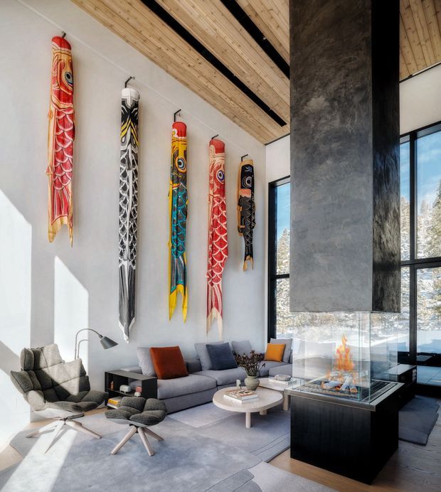 A spacious, contemporary living room with very high ceilings, exposed warm timber ceiling boards and dark structural beams. A dramatic tall concrete fireplace surround with a glass-enclosed fire anchors the room beside full-height black-framed windows. The space includes a grey sectional sofa, sculptural lounge chair and ottoman, low round coffee table, and a striking collection of vertically mounted colourful carved wall sculptures. Natural light, pale flooring and layered neutral furnishings create a refined, warm atmosphere.