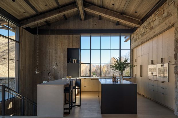 A warm, contemporary kitchen inside a high-ceilinged, timber-lined space with exposed structural beams and a rustic stone wall. Two dark kitchen islands with integrated seating face a large black-framed window overlooking a dry, mountainous landscape. Pale wood cabinetry, dark benchtops, pendant lights, and expansive glazing create a refined rustic-modern atmosphere.