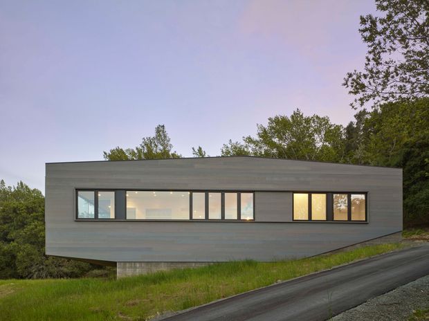 Exterior view of a contemporary single-storey rectangular house set on a sloping, wooded site. The building has a long, low-profile form with dark grey horizontal cladding, a shallow roofline, and a continuous band of narrow rectangular windows, some glowing warmly from the interior. The structure appears slightly elevated above the grassy slope, with a paved driveway in the foreground and mature trees surrounding the property.