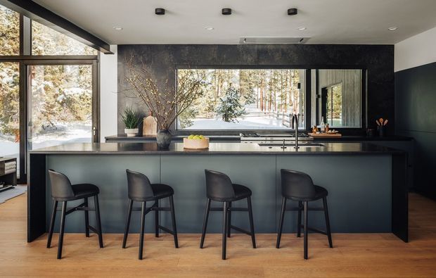 A modern kitchen with a long dark island and four bar stools, dark cabinetry, a black stone-look splashback, timber flooring, and expansive timber-framed glazing overlooking a snowy wooded landscape. The kitchen has a clean, sophisticated fit-out with recessed ceiling lights and a large horizontal window above the benchtop.