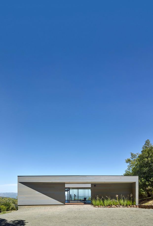 A minimalist single-storey contemporary house viewed from the front, set on a landscaped sloping site. The building has a simple flat-roofed rectangular form with horizontal light timber cladding, a large recessed central glazed opening, and a deep covered outdoor area. A gravel forecourt, native-style planting, trees, and distant hills surround the structure beneath a clear blue sky.