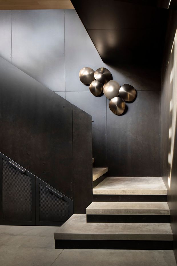 A dramatic modern interior stairway with broad concrete steps, dark metal wall and stair balustrade panels, and a black ceiling plane. A sculptural cluster of metallic spherical wall art is mounted above the stairs, while narrow daylight enters from the right through vertical glazing or a slatted opening. The space has a refined industrial character with strong geometric lines and high contrast.