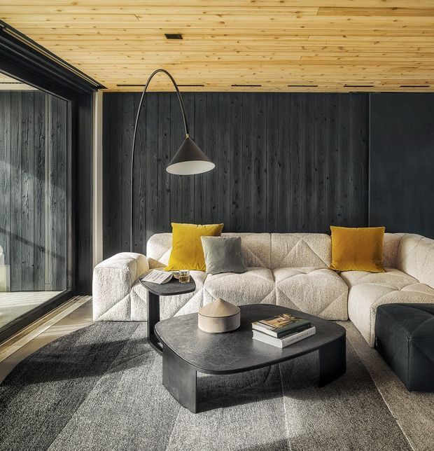 A refined living room with a large cream tufted sectional sofa, dark vertical timber wall panelling, and a warm timber-lined ceiling. A black sculptural coffee table, charcoal rug, arched pendant light, and mustard and grey cushions create a modern, high-contrast interior. Full-height glazed doors appear at the left, bringing in natural light.