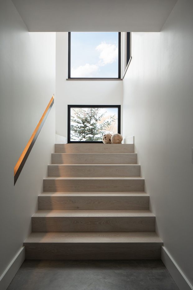 A minimalist stairway viewed from below, with broad light timber treads and risers ascending between smooth white walls. A dark grey concrete-like floor anchors the foreground, while slim black-framed windows bring in daylight and views of trees and sky. A simple timber handrail with a dark support runs along the left wall, and small decorative objects sit on the upper landing.