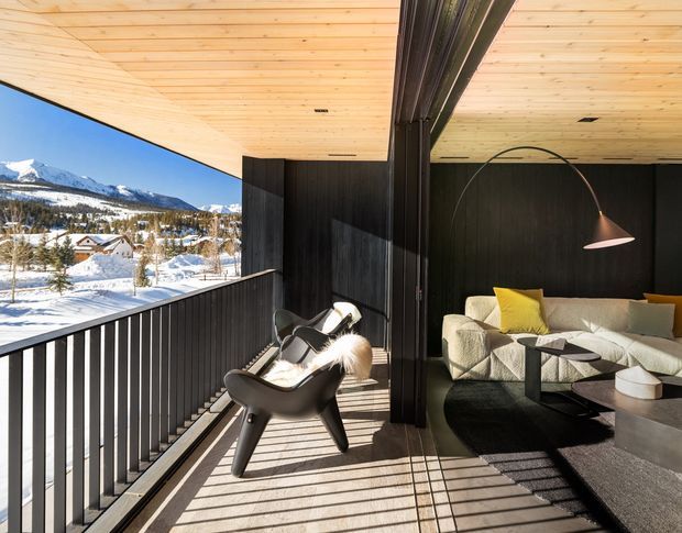 A sophisticated mountain-view living area opening onto a covered balcony or terrace. The space features black vertical wall cladding, a warm timber-lined ceiling, dark flooring, sculptural lounge chairs, a light modular sofa with yellow accent cushions, a low black coffee table, and a curved floor lamp. Snow-covered mountains and a residential landscape are visible beyond the black metal balustrade.