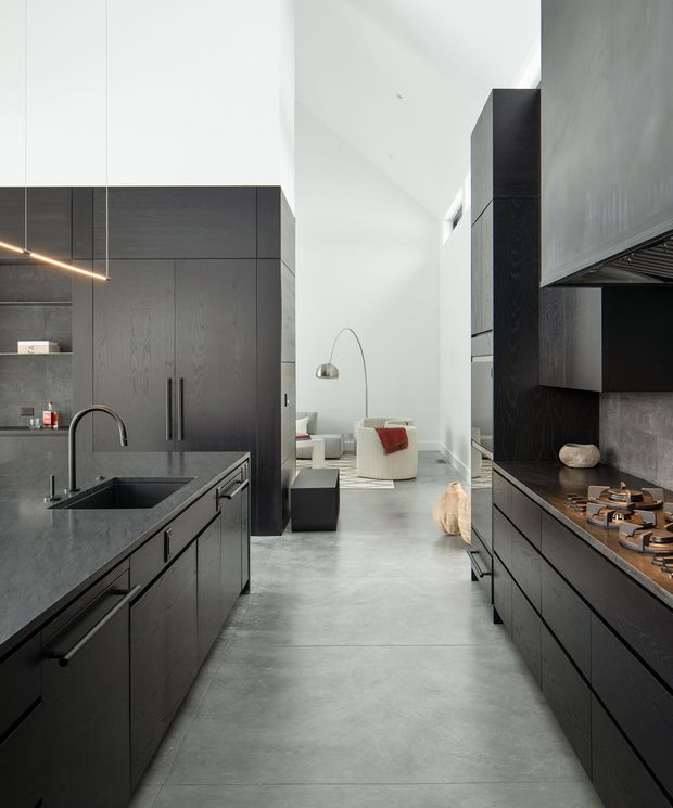 A contemporary galley-style kitchen with dark timber-look cabinetry on both sides, a large island with an integrated black sink and curved tap, and a cooking run with a timber-toned benchtop and gas cooktop. Pale concrete-look flooring and white walls create contrast, while a tall open living area is visible beyond the kitchen. The space has a restrained, high-end minimalist aesthetic with linear pendant lighting and concealed integrated appliances.