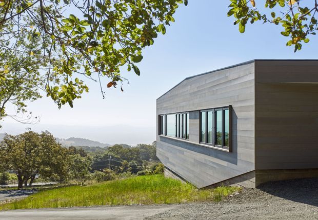 Exterior view of a contemporary single-storey building on a sloping, elevated site. The structure has a low-pitched asymmetrical roofline, horizontal grey timber-like cladding, a dark side wall, and a long band of black-framed windows. A gravel driveway and landscaped grassy surroundings are visible, with a distant wooded valley beyond.