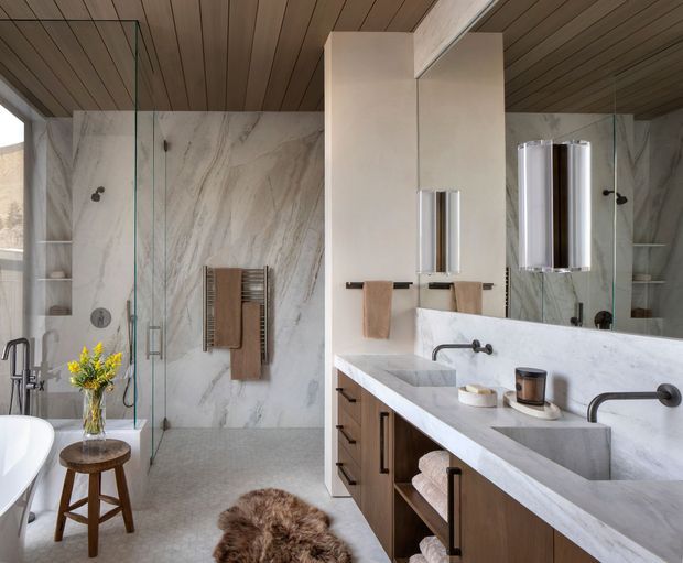 Luxurious contemporary bathroom with a large walk-in glass shower, freestanding bathtub, double vanity, and full-height marble-look wall surfaces. Warm timber ceiling boards and timber vanity cabinetry contrast with the pale stone finishes. A heated towel rail, wall-mounted fittings, mirrors, and understated accessories complete the refined, spa-like space.