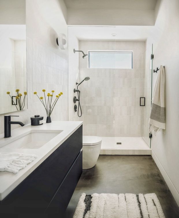 Bright contemporary bathroom with a walk-in shower enclosed in pale tiled walls, a high horizontal window, wall-mounted shower fittings, and a white toilet. In the foreground is a large floating dark vanity with a white integrated basin and matte black tapware. A mirror, simple decorative stems, towel hooks, and a textured bath mat complete the clean monochrome scheme.