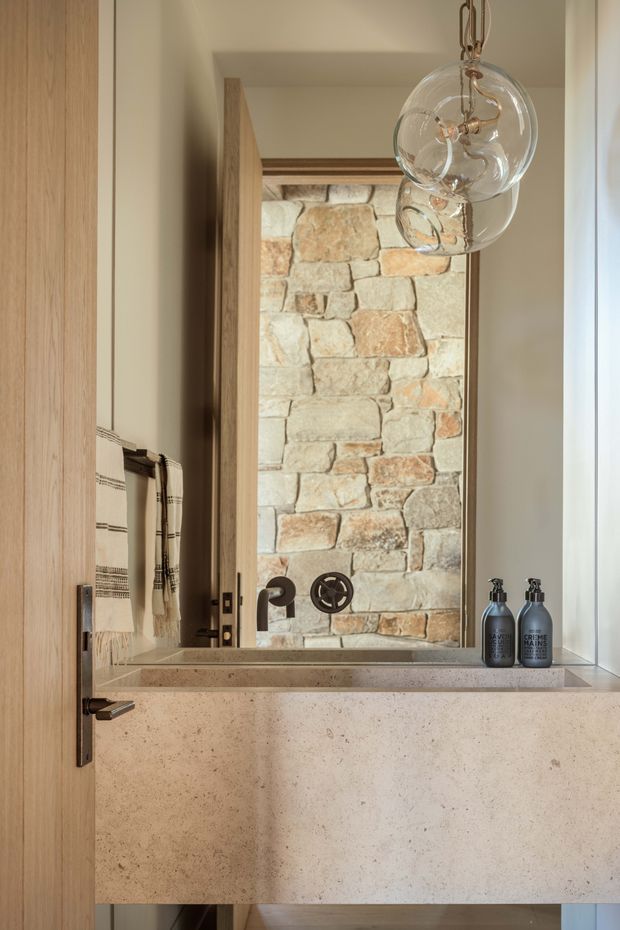 A refined bathroom vanity viewed through an open timber door. The room features a thick pale stone or terrazzo-like basin and benchtop, a large mirror framed by exposed irregular stone masonry, dark bronze wall-mounted tapware, timber joinery, and clear globe pendant lights. Neutral warm tones and natural materials create a calm, contemporary-rustic atmosphere.