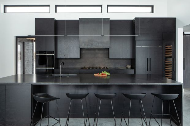 A sleek, contemporary kitchen dominated by a large dark island with five black bar stools. The kitchen features matte dark cabinetry, integrated appliances, a broad dark benchtop, a central stainless-steel rangehood, and a dark tiled or stone-look splashback. Narrow clerestory windows provide daylight above the cabinetry, while an opening to the side reveals an adjacent area.
