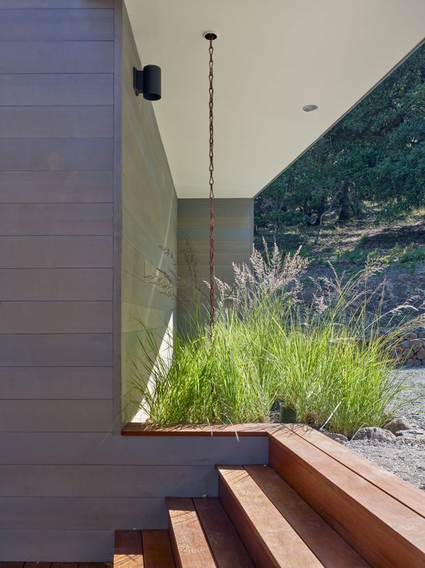A sheltered exterior entry or passage beside a contemporary building, featuring horizontal timber-clad walls, a flat pale ceiling with recessed lighting, a black wall sconce, and a suspended chain-like pendant. Broad timber steps and bench-like decking lead through a planted threshold with tall ornamental grasses, opening toward a rocky, wooded landscape.