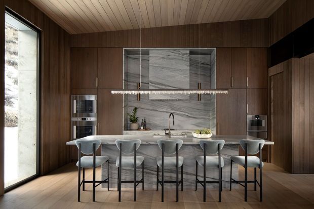 A sophisticated contemporary kitchen with a long marble island and five bar stools, dark timber cabinetry and wall panelling, a dramatic grey-veined stone splashback, integrated appliances, and a linear pendant light. Full-height glazing at left brings in natural light, while the timber-lined ceiling and warm wood floor create a refined, intimate atmosphere.