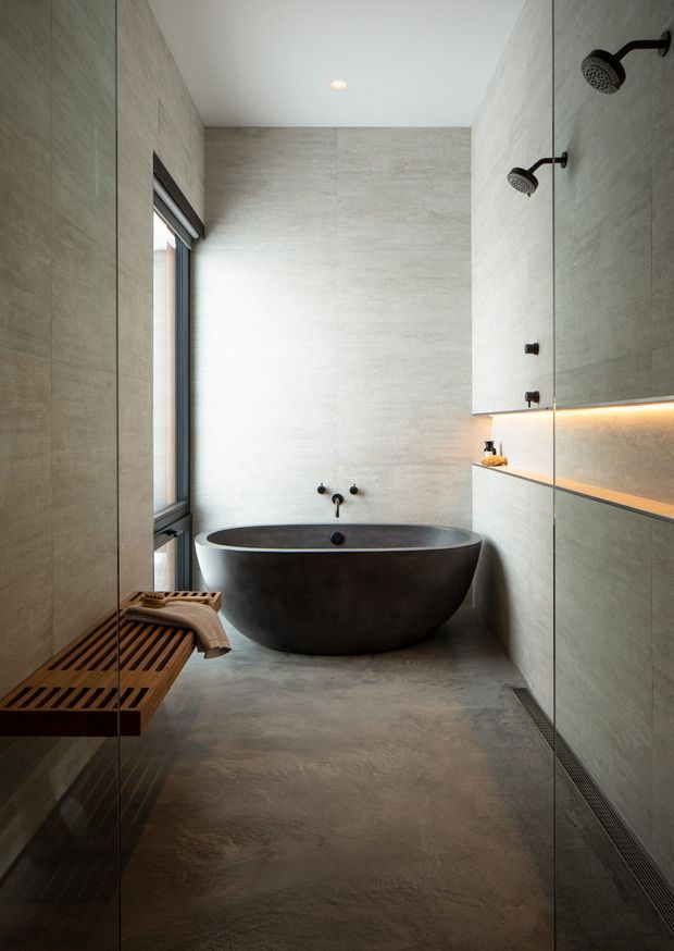 A refined contemporary bathroom with a freestanding dark oval bathtub positioned against large-format pale grey stone-look wall and floor surfaces. A glass-enclosed walk-in shower occupies the right side, with twin dark showerheads, recessed fixtures and warm linear under-cabinet lighting. A narrow window, timber slatted bench and folded towel add natural warmth, while a long linear floor drain and minimalist fittings reinforce the clean design.