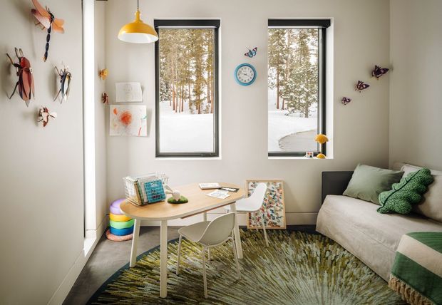 A bright, playful children's room with two tall windows overlooking a snowy wooded landscape. The room contains a compact light-wood desk with white chairs, a sofa/daybed with green cushions and a knitted green throw, a colourful textured rug, children's artwork, and whimsical insect and butterfly wall decorations. The palette is predominantly white, cream, light wood, and green with colourful accents.