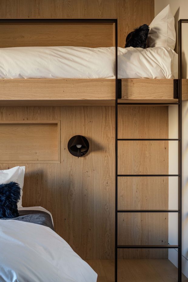 A compact bedroom featuring a custom built-in timber bunk-bed and storage wall. Warm wood panelling and integrated shelving are framed with slim black metal rails, with two neatly made beds, white bedding, dark cushions, and a black wall light creating a refined, space-efficient design.