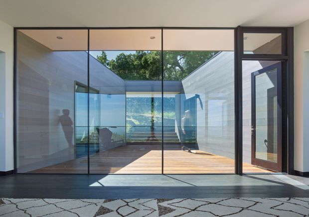 A bright contemporary interior looking through full-height glazed doors into a landscaped courtyard or terrace. The space features dark timber or metal-framed glazing, pale walls, a warm timber deck, and an open-air central court with steps and greenery beyond. Reflections and silhouettes are visible in the glass, while a patterned rug occupies the foreground.