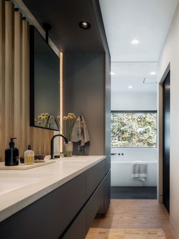 A contemporary bathroom with a long double vanity, integrated white benchtop and undermount basins, matte dark cabinetry, warm vertical timber wall panelling, and a large mirror. A black curved tap, recessed ceiling lights, towels, and decorative vessels add refined detail. The vanity leads toward a brighter bathing area with a large window and towel rail, while light timber flooring warms the space.