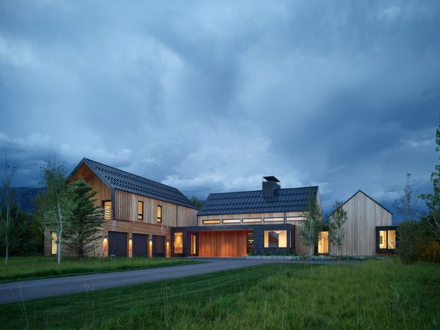 Exterior architectural rendering of a contemporary multi-building residence in a landscaped rural or semi-rural setting. The composition shows several connected and detached timber-clad volumes with steep pitched metal roofs, warm illuminated windows, a central entry or garage-like opening, and a dark stormy sky. A driveway or paved approach runs across the foreground.