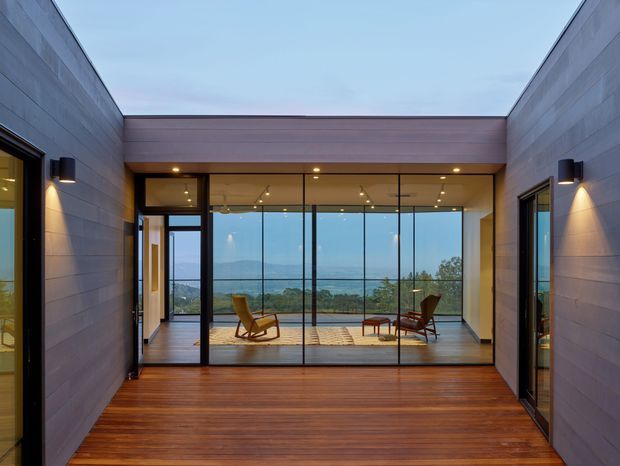 A modern courtyard-like outdoor passage with dark timber decking, grey horizontal wall cladding, and concealed wall lighting leads to a fully glazed living area. Through the floor-to-ceiling glass, two lounge chairs and a low table are visible, with expansive water and landscape views beyond. The image is photographed from outside, emphasizing the contemporary building envelope, glazing, and connection between the courtyard and interior.