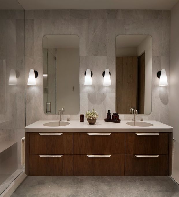 A refined contemporary bathroom centered on a wide double-sink vanity with dark timber drawer cabinetry and a white solid benchtop. Two rounded-rectangle mirrors sit above the basins, flanked by sculptural wall sconces. Large pale stone-look wall tiles, a grey floor, glass shower enclosure, and warm wood tones create a calm, softly lit interior.