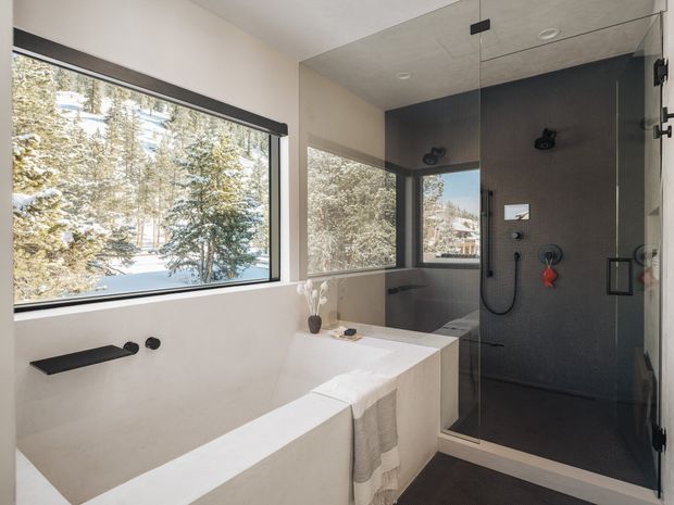 Contemporary bathroom with a freestanding white bathtub beside a fully glazed walk-in shower. The shower has dark charcoal wall tiling, black fixtures and fittings, and a frameless glass enclosure. Large windows provide views of a wooded, snowy landscape, while pale walls and white sanitaryware create a bright, clean interior.
