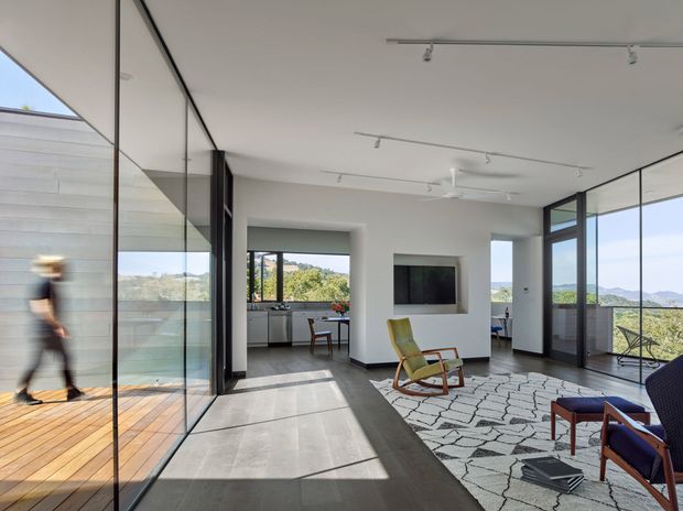Bright contemporary open-plan living space with expansive floor-to-ceiling glazing, polished concrete flooring, and a white minimalist interior. A glazed terrace or balcony runs along the left side, while the living area contains a patterned rug, mid-century-style lounge chairs, a wall-mounted television, and track lighting. Views of a green, hilly landscape are visible through the windows.