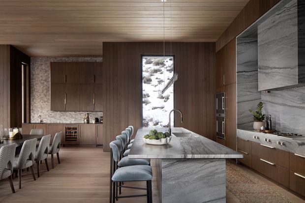 A sophisticated open-plan kitchen and dining area with extensive dark timber cabinetry, a large grey-veined stone island and matching splashback, integrated appliances, and a timber-lined ceiling. The island includes seating for four, while a dining table with upholstered chairs sits to the left. A tall window brings in a view of a snowy, rocky landscape. The room has a warm, refined contemporary mountain-lodge character.