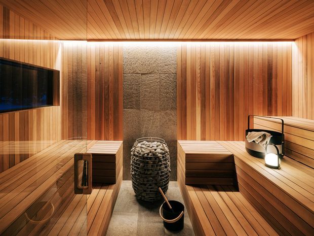 A warm, compact sauna clad throughout in vertical and horizontal timber slats, with tiered bench seating on both sides, a central stone-clad sauna heater filled with rocks, and soft concealed lighting. A dark glazed panel or screen is visible on the left, while sauna accessories sit on the right bench.