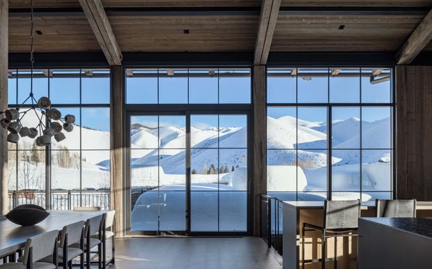 A dramatic open-plan interior with exposed timber beams and dark timber ceiling, floor-to-ceiling black-framed glazing, and a contemporary dining and kitchen area. The glazing provides expansive views of snow-covered mountains and a winter landscape. A sculptural pendant light hangs above the dining table, while dark cabinetry and streamlined furnishings create a refined alpine retreat atmosphere.