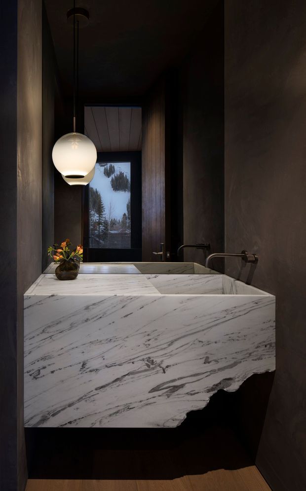 A dark, luxurious bathroom viewed toward a sculptural wall-mounted double vanity. The vanity is clad in heavily veined white natural stone with an irregular live-edge lower profile, paired with integrated rectangular basins and wall-mounted dark metal tapware. Deep brown or charcoal walls and vertical timber panelling create a moody atmosphere, while a spherical pendant light illuminates the marble surface. A window at the rear reveals a wooded, snowy landscape.