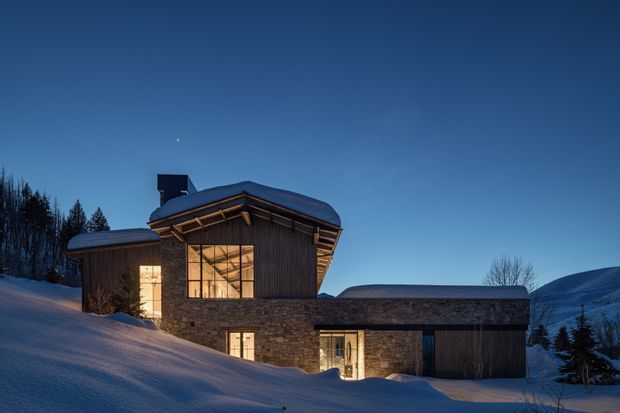 Exterior view of a contemporary mountain house at dusk, partially embedded into a snowy sloped site. The building combines dark vertical cladding, extensive warm-lit glazing, and textured stone walls beneath a low, overhanging pitched roof with exposed timber eaves. Snow-covered ground, trees, and surrounding alpine terrain frame the residence.