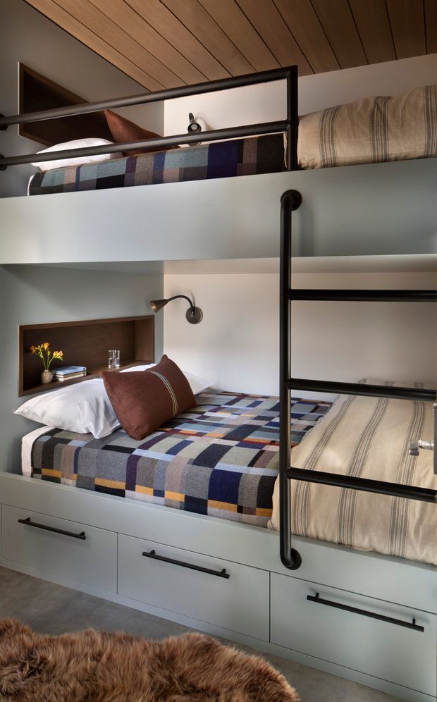 A compact, built-in bunk-bed room with two stacked beds, integrated drawers beneath the lower bunk, dark metal safety rails and ladder, and recessed bedside niches with wall-mounted reading lights. Both bunks are dressed in patterned multicolour blankets and neutral striped bedding. The room has a warm timber-lined ceiling, pale blue-grey built-in joinery and walls, dark hardware, and a partial shaggy brown rug.