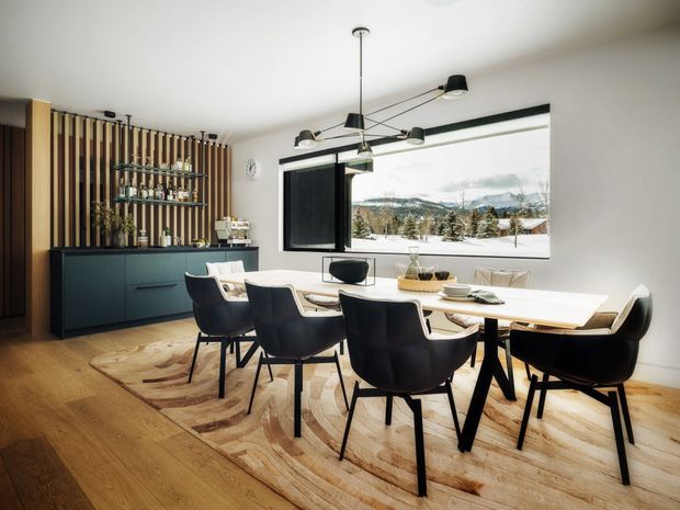 A contemporary dining room with a long white dining table surrounded by dark upholstered chairs, set on a large neutral patterned rug over warm timber flooring. A dark built-in sideboard and open shelving with glassware sit against a vertical timber-slat feature wall. Large black-framed glazing provides a mountain view, while a sculptural multi-arm pendant light hangs above the table.