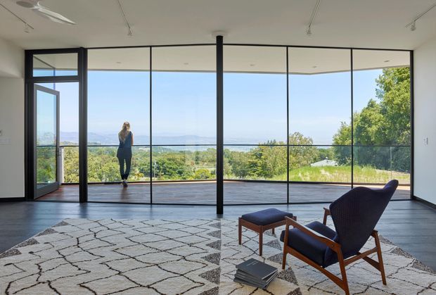 A spacious, minimalist living room with dark timber flooring, white walls and ceiling, and extensive black-framed floor-to-ceiling glazing opening onto a timber deck. The room contains a patterned area rug, a mid-century-style lounge chair with ottoman, and a small stack of books. The glazing provides expansive views over a green landscape and distant water or hills, creating a bright, airy connection to the outdoors.
