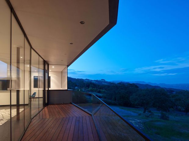 A modern, elevated terrace at dusk with broad timber decking, frameless glass balustrades, and floor-to-ceiling glazing opening onto expansive rolling bushland and distant hills. The cantilevered roof soffit and recessed downlights create a sheltered outdoor living edge, while the interior workspace is visible through the glazing.