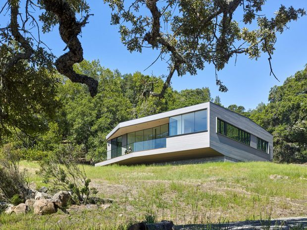 A low-slung contemporary house set into a wooded, gently sloping landscape. The building has a strong horizontal form, a shallow angular roofline, extensive floor-to-ceiling glazing, and horizontal grey exterior cladding. Mature trees and natural rocky terrain surround the residence.
