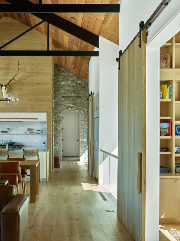 A bright, vertically composed residential interior showing a hallway opening into a dining and kitchen area. The space features pale timber flooring, a warm timber-lined pitched ceiling, exposed dark structural beams, a substantial natural stacked-stone feature wall, and large sliding barn-style timber doors. Built-in timber shelving is visible on the right, while the dining table, leather chairs, kitchen cabinetry and island appear to the left. The design has a warm, contemporary-rustic character with strong natural materials and abundant daylight.
