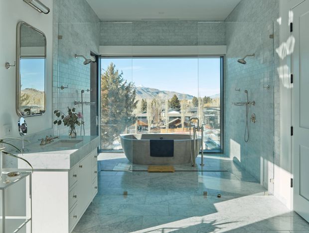 A bright, spacious contemporary bathroom with pale blue-grey marble-look wall and floor surfaces, a freestanding bathtub positioned in front of full-height glazing, a glass-enclosed shower area, and a long white vanity with integrated basin and drawers. Large windows overlook trees, rooftops, and mountainous terrain. Chrome fixtures, a frameless shower enclosure, and strong natural sunlight create an airy, refined atmosphere.