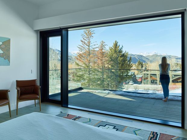 A bright contemporary bedroom with a large black-framed glazed opening overlooking a timber terrace, snow-capped mountain landscape, and trees. The room has pale walls, a low bed in the foreground, leather lounge chairs, and a geometric patterned rug. A person stands outside on the terrace, emphasizing the expansive indoor-outdoor connection.