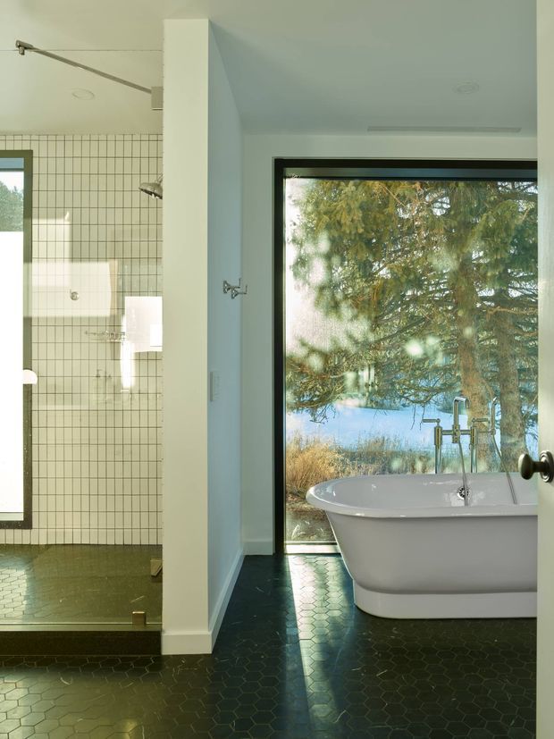 A bright contemporary bathroom with a freestanding white bathtub positioned beside a large black-framed window overlooking trees and water. The room has dark hexagonal floor tiles, a white tiled walk-in shower with a slim black grid pattern, pale walls, and minimalist chrome plumbing fixtures. A partial dividing wall separates the shower area from the bath.